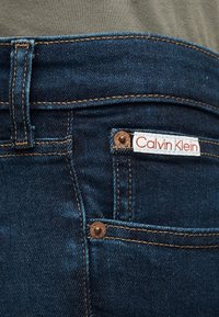 Τσέπη από σκουρόχρωμο μπλε τζιν με χάλκινα καρφιά και λευκή ετικέτα Calvin Klein ραμμένη στο πάνω άκρο της τσέπης.