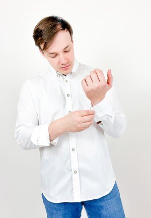 Homme ajustant un bouton de manchette sur une chemise blanche à boutons, debout devant un fond clair uni.