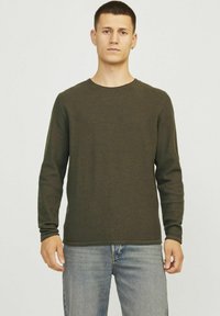 Pulover texturat cu maneci lungi, de culoare verde închis, cu guler rotund, asortat cu blugi din denim de culoare albastră deschisă. Materialul pare a fi cu model în falduri și are o siluetă ajustată.