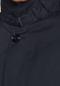 Černá, texturovaná tkanina s vysokým límcem, vybavená uzávěrem na knoflíky a nastavitelnými detaily na popruzích. Klasický design s minimálním zdobením.