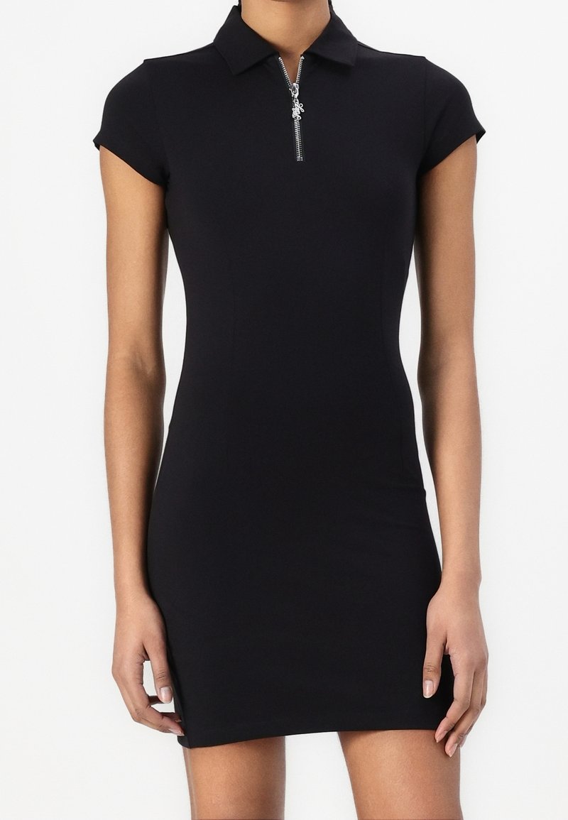 Robe noire cintrée à manches courtes avec col et fermeture éclair argentée à l’avant, portée par une personne debout, montrée du cou jusqu’au milieu de la cuisse.