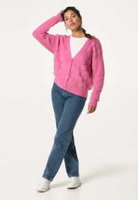 Roze gebreide cardigan met een gestructureerd bubbelpatroon, knoopsluiting en verlaagde schouders, gecombineerd met blauwe jeans en witte sneakers.