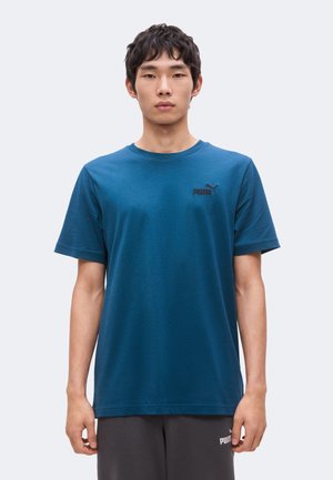 Giovane uomo con capelli corti e scuri che indossa una maglietta blu Puma e pantaloni Puma scuri, in piedi davanti a uno sfondo chiaro e uniforme.