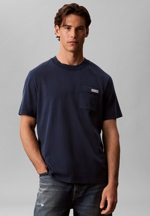 Uomo che indossa una t-shirt blu scuro a maniche corte con taschino sul petto e jeans blu sbiaditi, in piedi davanti a uno sfondo grigio uniforme.