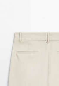 Back view of beige pants with belt loops and two welt pockets on a white background.