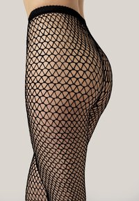 Nærbillede af en persons underkrop iført sorte netstrømpebukser med et diamantmønster mod en neutral baggrund.