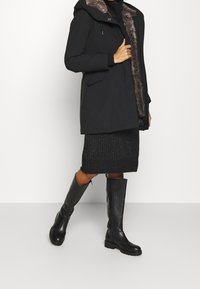 Svart parkacoat med ett pälsfoder på kragen, två framfickor, lager på en svart stickad kjol och tillsammans med knähöga svarta läderstövlar.