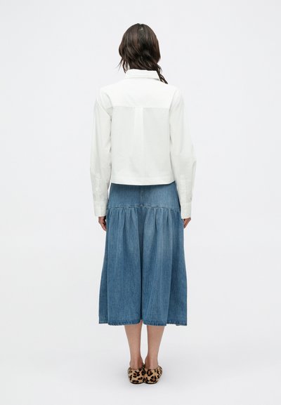 Biela skrátená košeľa s dlhými rukávmi, doplnená modrou džínsovou sukňou s vrstvami. Na spodku sú viditeľné semišové mokasíny s leopardím vzorom.