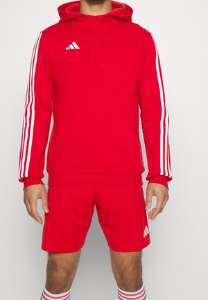Hombre con sudadera con capucha roja de Adidas con rayas blancas en las mangas y pantalones cortos rojos a juego con el logo de Adidas en la pierna izquierda.