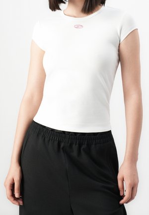 Femme portant un t-shirt court blanc ajusté avec un petit logo rouge au niveau de l'encolure et un pantalon noir ample avec une taille élastique.