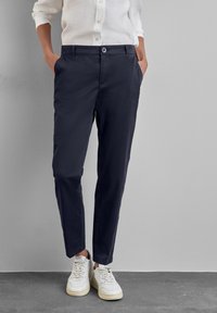 Henkilö pukeutuneena tummansinisiin räätälöityihin housuihin, valkoiseen kauluspaitaan ja valkoisiin lenkkareihin, seisomassa kädet taskuissa harmaan seinän edessä.