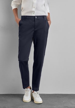 Person trägt marineblaue, maßgeschneiderte Hose, weißes Hemd mit Knöpfen und weiße Turnschuhe und steht mit den Händen in den Taschen vor einer grauen Wand.