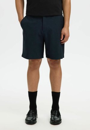 Homme portant des mocassins en cuir noir, des chaussettes noires mi-mollet, un short bleu marine foncé jusqu'aux genoux et une chemise noire, les mains dans les poches.