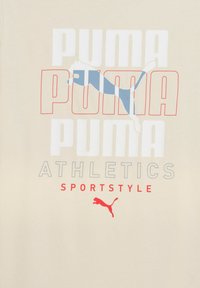 Beige t-shirt med gentaget "PUMA" tekst i rød og blå, med en puma silhuet. Designet fremhæver atletiske og sportslige elementer.