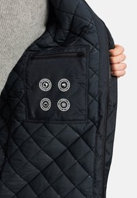 Den svarte, quiltede jakkeinnsiden har en glidelåslomme med fire sirkulære ikoner som indikerer vannavstøtende, vindtett, varme og kulderesistens.