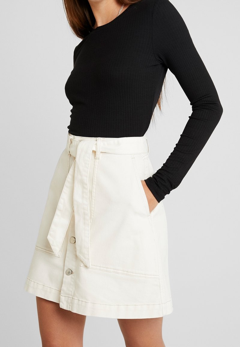 Vithe denimkjol med sidofickor, knytskärp i midjan och framsidans knappstängning. En ribbad svart långärmad topp kompletterar outfiten.