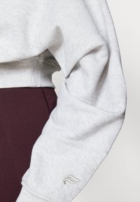 Lys grå sweatshirt med ribbede mansjetter, som har en løs passform. Har en liten brodert logo i sølv på ermene. Dype burgunder leggings synlige.