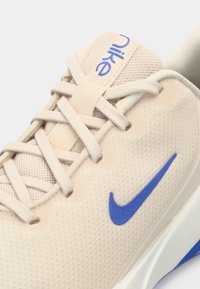 Helle beige Nike Sneakers mit blauem Logo, ausgestattet mit einem Obermaterial aus Mesh, flachen Schnürsenkeln und einer strukturierten Sohle. Nahaufnahme der Schuh-Design-Details.
