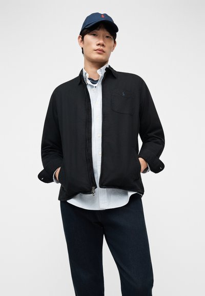 Νέος άνδρας που φοράει σκουρό μπλε καπέλο μπέιζμπολ, μαύρο μπουφάν πάνω από άσπρο πουκάμισο με ρίγες και σκουρό μπλε παντελόνι, στέκεται με τα χέρια στις τσέπες.