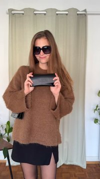 Καφέ χνουδωτό πουλόβερ, μαύρο clutch Moschino, μαύρη φούστα με σκίσιμο, υπερμεγέθη μαύρα γυαλιά ηλίου, στέκεται μπροστά σε ανοιχτοπράσινη κουρτίνα.