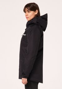 Veste isolée noire avec capuche, fermeture éclair à l'avant, poches latérales et poignets ajustables. Texture lisse et minimalisme en matière de branding.