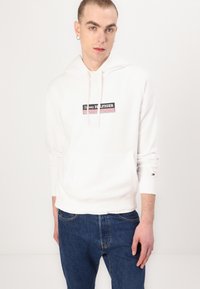 Hvid bomulds hoodie med en frontlomme, snøre-hætte og et trykt logopanel i sort og rød. Bæres med blå jeans.
