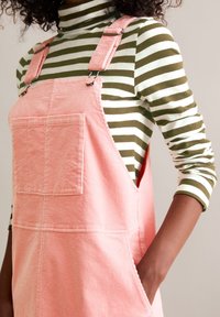 Person in rosa Kordoverall mit Fronttasche über einem langärmeligen grün-weiß gestreiften Shirt, Hand in der Seitentasche.