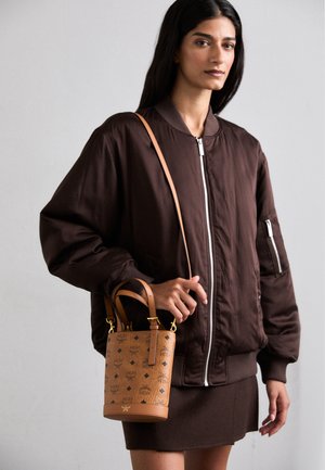 Bruine bomberjack met een rits, gecombineerd met een tan kleurige bucket bag met logo's en een gouden sluiting. De tas heeft korte handvatten en een lange schouderband.