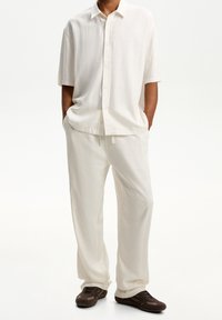 Camisa blanca de lino de manga corta con botones en el frente, combinada con pantalones de lino blancos de corte suelto, usados con zapatillas marrones.