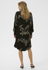 Plava žena koja nosi crnu midi haljinu s zlatnim cvjetnim paisley uzorcima, dugim rukavima i crnim sandalama s mačkicom, stoji okrenuta leđima.