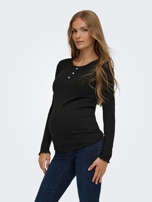 Femme enceinte aux longs cheveux portant un haut noir à manches longues avec des boutons et un jean bleu foncé, debout avec une main sur la hanche.
