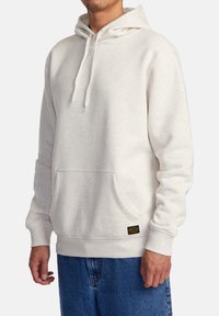 Un sweat à capuche gris clair avec des cordons de serrage ajustables, une poche frontale, des poignets côtelés et un patch avec logo en bas à gauche. Texture de tissu douce.