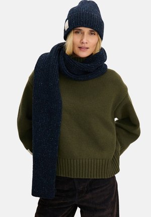 Kobieta w ciemnoniebieskiej dzianinowej czapce i pasującym długim szaliku, ubrana w oliwkowy sweter i ciemne spodnie na białym tle.