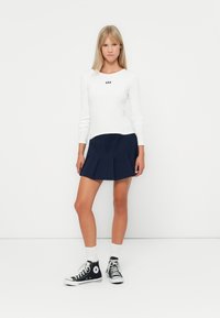 Witte geribbelde longsleeve shirt met het "GAP" logo, gecombineerd met een marineblauwe geplooide skort. Het model draagt zwarte hoge sneakers met witte sokken.