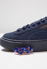 Marineblaue Puma Wildlederschuh mit dicker schwarzer Sohle, Platzierung eines blauen Edelsteinbroches mit rosévergoldetem Rahmen auf weißer Oberfläche.