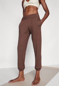 Brązowe spodnie sportowe z elastycznym pasem, bocznymi kieszeniami i ściągaczami u dołu. Posiadają subtelne czarne logo na udzie.