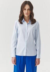 Camisa azul claro de botones en tejido suave, con mangas largas, cuello puntiagudo y cierre de botón clásico.