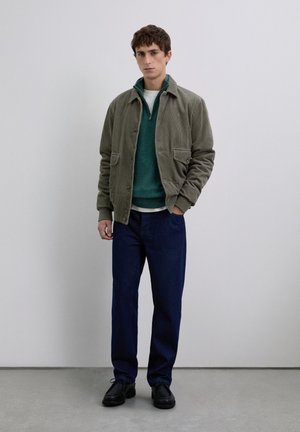 Jeune homme portant une veste en velours côtelé olive, un pull-over à zip teal, un t-shirt blanc, un pantalon bleu foncé et des chaussures noires, se tenant à l'intérieur.