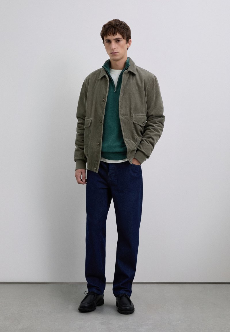 Jeune homme portant une veste en velours côtelé olive, un pull-over à zip teal, un t-shirt blanc, un pantalon bleu foncé et des chaussures noires, se tenant à l'intérieur.