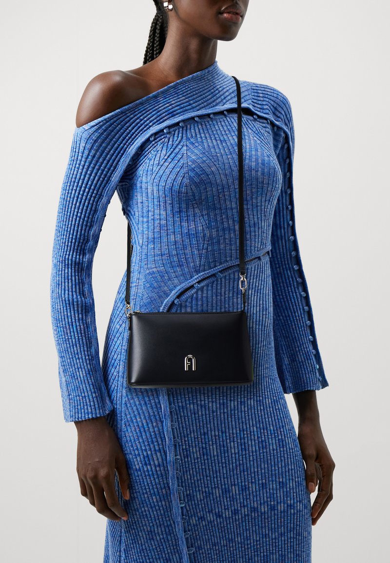 Borsa a tracolla in pelle nera con logo argentato, texture liscia e tracolla regolabile, indossata sopra un vestito in maglia blu a costine.