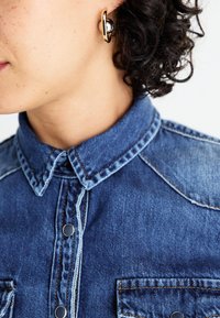 Camicia in denim con un lavaggio blu chiaro, dettagli di cucitura visibili e bottoni neri. Orecchino a cerchio dorato con una perlina nera.
