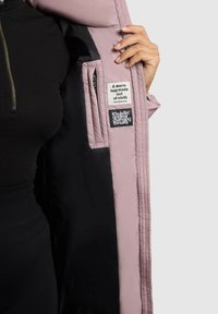 Giacca imbottita con un esterno rosa chiaro e fodera nera. Presenta etichette interne con testo, chiusura con cerniera e texture trapuntata.