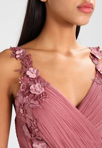 Laventelimekko, jossa on V-aukko, koristeltu brodeeratuilla kukkakuoseilla ja teksturoiduilla poimuilla yläosassa. Koristeina on helmikoristelua.