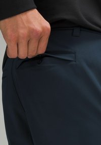 Mornarske hlače z gladko teksturo, ki imajo gumbasto stransko žep in prilegajoč dizajn za elegantno podobo. Roka je položena na žep.