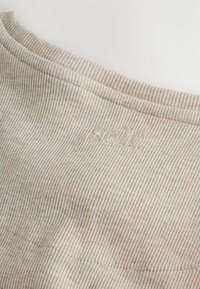 Beige bordázott anyag finom textúrával, amelyen egy kis hímzett logó található. Puha, rugalmas anyag, lekerekített nyakkivágással.
