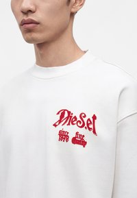 Jeune homme portant un sweat-shirt blanc avec un texte rouge brodé "Diesel 1978 fine quality" sur la poitrine.