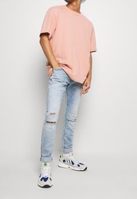 Camiseta de algodón rosa, de corte holgado. Jeans de mezclilla desgastados de color azul claro con roturas. Zapatillas blancas con acentos azules y verdes. Sombrero tipo bucket con ala plana.