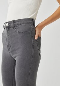 Jeans en denim gris taille haute avec une texture lisse, des poches avant et un accent en métal argenté au niveau de la taille.