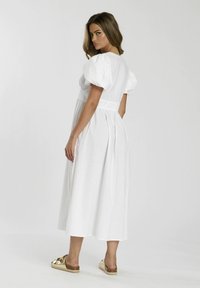 Weißes Midi-Kleid mit Puffärmeln, tailliertem Schnitt und gerafftem Rock. Das Model trägt goldene Sandalen mit Korksohle und metallischen Akzenten.