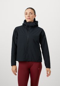 Črna zip-up jakna s kapuco, z gladko teksturo in nastavljivimi vrvicami. Nanjšena je nad rdečimi leginami, ima stranske žepke.
